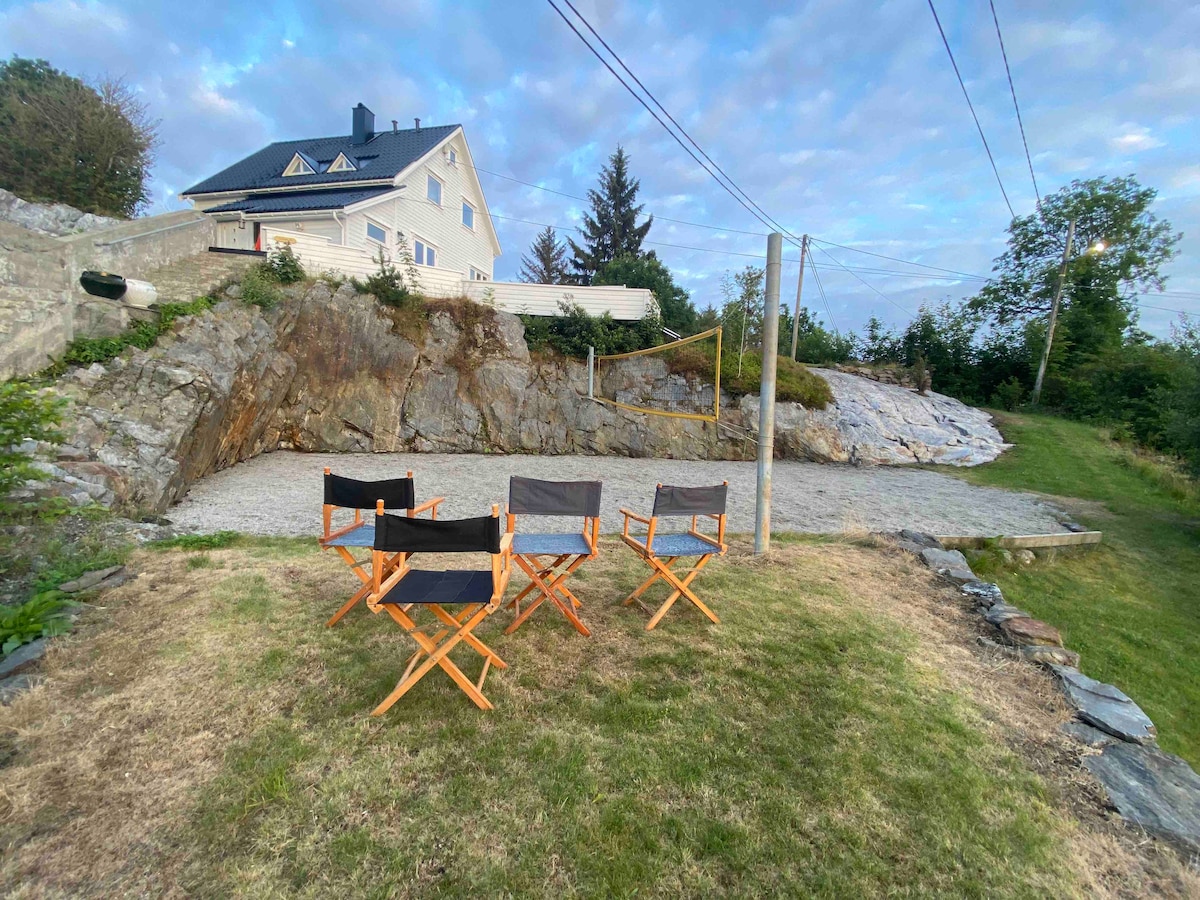 A gravel area is shown with four canvas chairs on wooden frames arranged in a semi-circle. The backdrop features a rocky landscape and green grass, with distant trees and a white house visible under a partly cloudy sky.