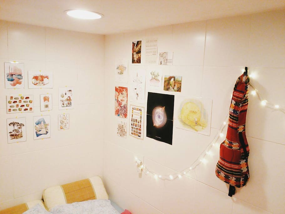 Cantinho da cama com tomada, luzinha de natal de cabeceira e muita arte na parede.