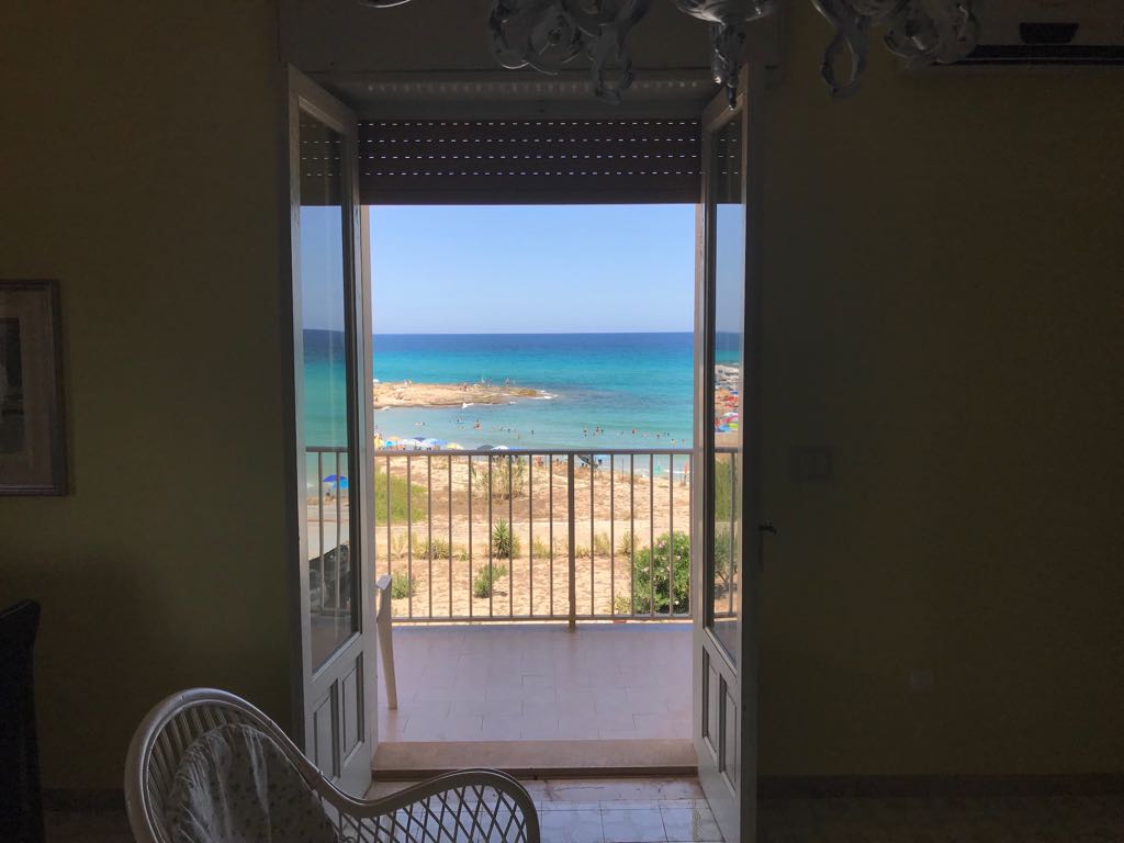 A view of the turquoise sea is framed by open double doors leading to a balcony. Gentle waves lap at the sandy beach below, with beachgoers enjoying the sun. Natural light fills the space, highlighting the serene coastal setting.