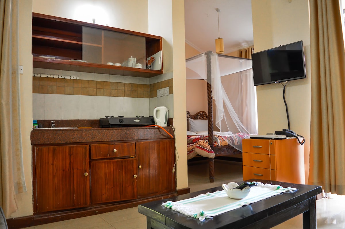A kitchenette is shown with wooden cabinetry and a countertop, including a kettle and dishware. A cozy sleeping area is partially visible in the background, featuring a bed with a decorative canopy and colorful bedding. A small table and a television are also present.