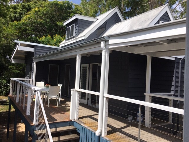 The elevated cottage features a covered verandah with a dining table and chairs positioned on a wooden deck. The exterior showcases a blue-grey facade, complemented by a gabled roof and large windows. Lush greenery surrounds the property, enhancing its tranquil setting.