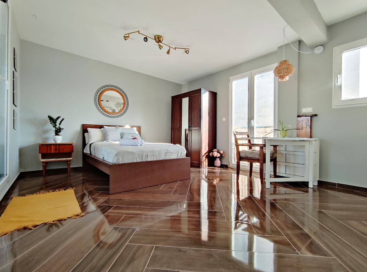 An open-concept studio with polished tile flooring features a spacious bed, a wall-mounted mirror, and a small dining area with a white table and chair. Light floods through large windows, complementing the neutral-colored walls and warm accents from decorative plants.