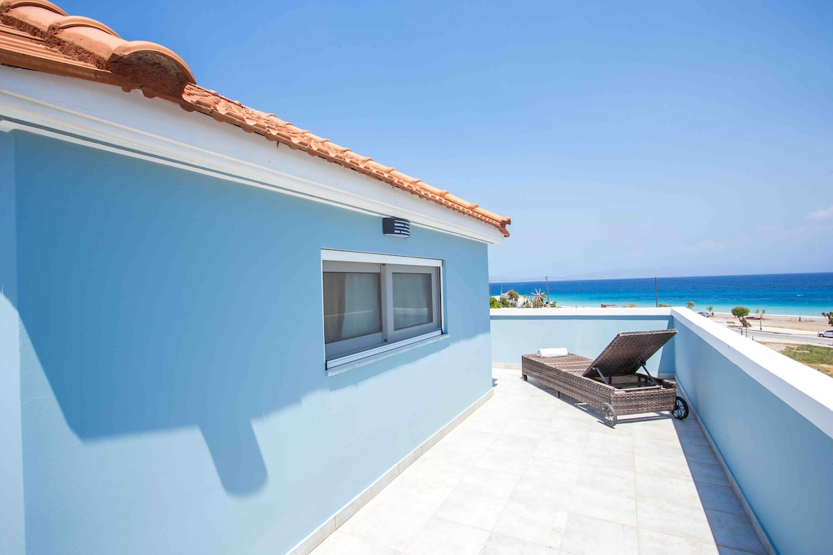 A spacious terrace features a relaxing lounge chair facing the sea, showcasing a clear blue sky above the tranquil waters. A minimalistic design is highlighted by smooth tiled flooring and the gentle curvature of the building's outline.