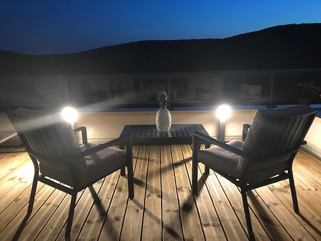 A spacious terrace features two chairs positioned around a small table, illuminated by two soft light sources. The wooden deck contrasts with the evening sky, and a decorative centerpiece adds a touch of elegance. The surrounding hills can be seen in the background.