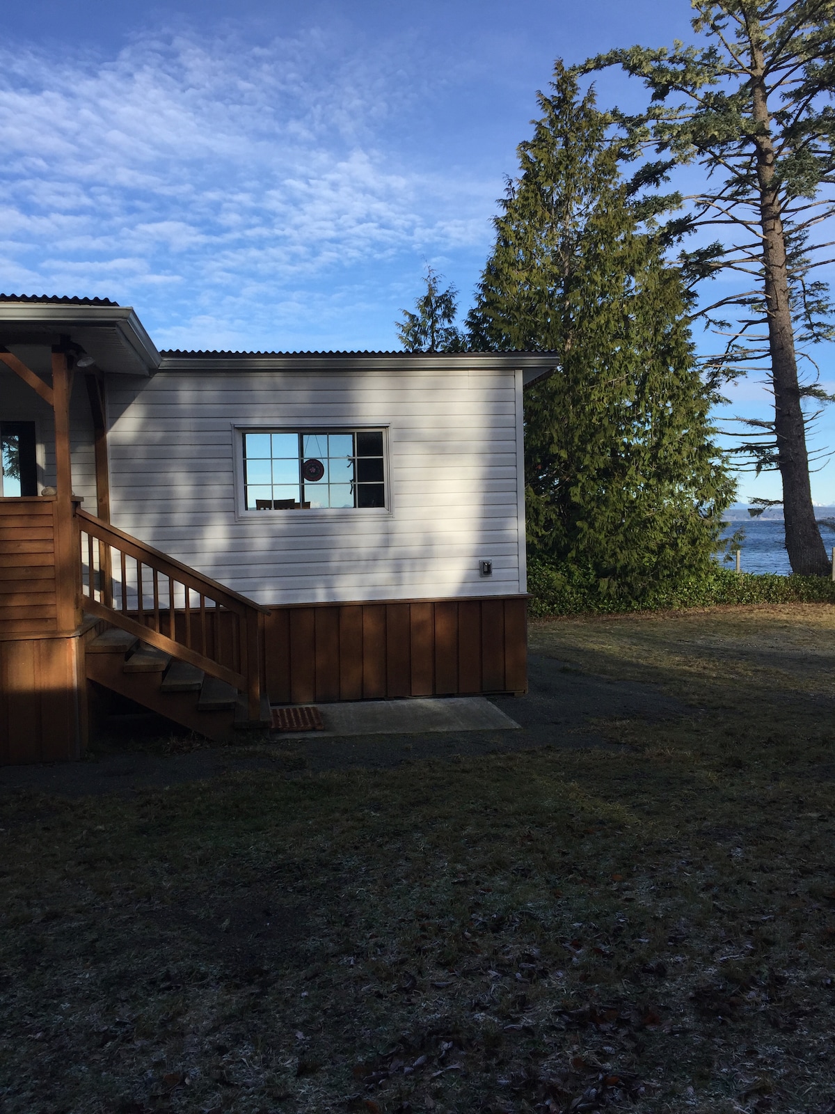 The exterior of a mobile home is depicted, featuring a wooden staircase leading to the entrance. A window is visible on the side, providing a glimpse of the ocean nearby. Tall trees frame the scene, with clear skies and an expansive green lawn.