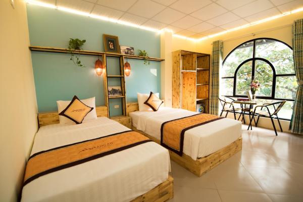 This inviting guest room features two spacious beds with natural wood frames. A decorative accent wall in soft blue sets a serene tone. Large windows allow natural light to illuminate a small dining area with a table and two chairs, overlooking the garden.