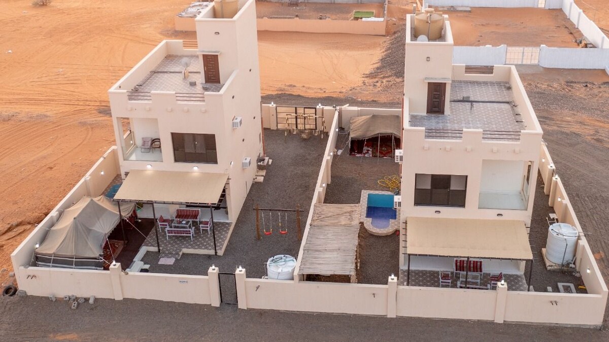 Two separate buildings are depicted, enclosed within a wall. Outdoor shaded areas feature seating, while a small swimming pool is visible. Swing sets are arranged nearby, and the desert landscape surrounds the compound, offering a clear view of neighboring sand dunes.