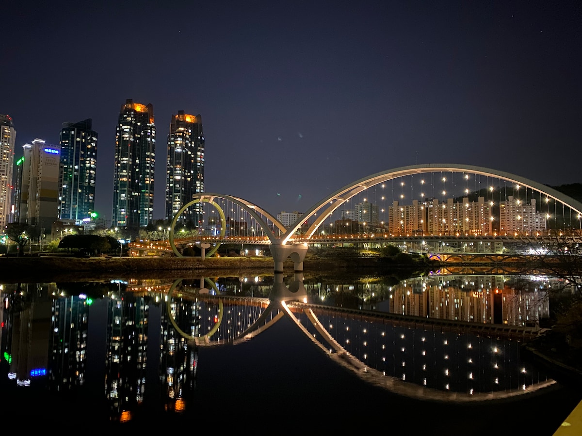 An urban scene is depicted at night, featuring illuminated modern skyscrapers alongside a bridge's striking arch. The reflection of the bridge and buildings is visible on the calm water below, creating a symmetrical visual effect with soft lighting enhancing the setting.