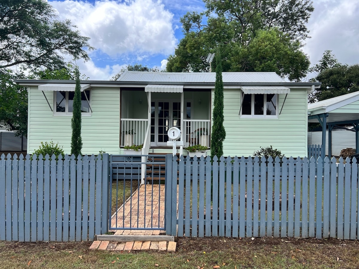 A light green retro Queenslander cottage is framed by a pale blue picket fence and a wooden pathway leading to the entrance. Tall, slender cypress trees flank the front steps, and the home features a covered porch with steps leading to the yard, illuminated by natural daylight.