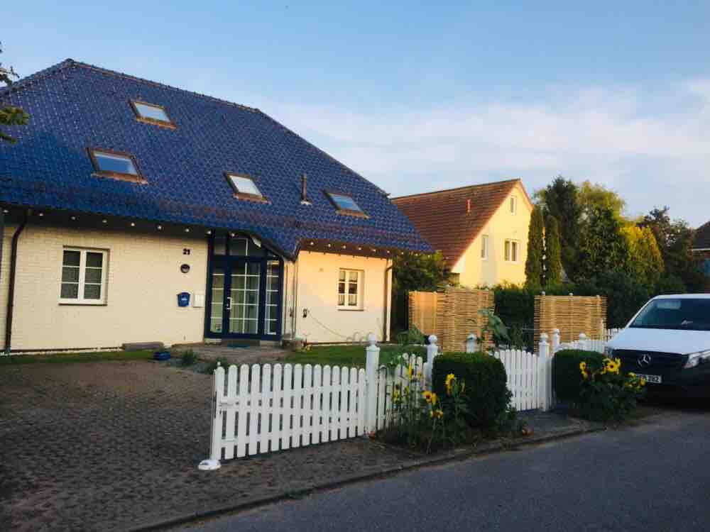 A modern two-story building features a striking blue roof and large front windows. Decorative outdoor lighting enhances the façade. A white picket fence encloses a garden area adorned with sunflowers, complementing the surrounding greenery and landscaped yard.