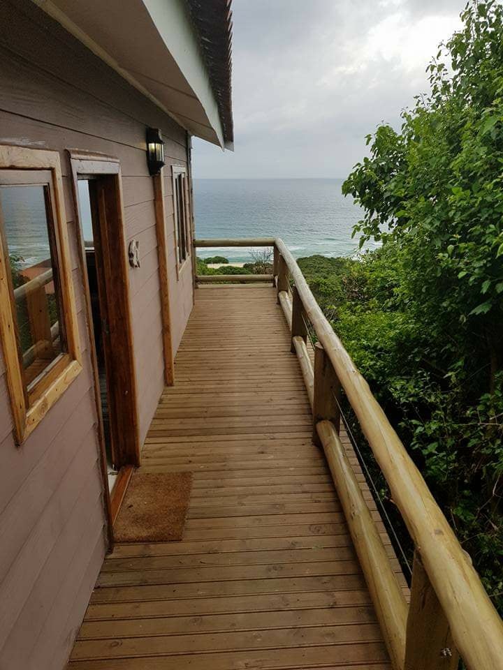 A wooden balcony extends from the exterior, offering unobstructed views of the ocean. The railing, made of natural wood, complements the surrounding greenery. Two doors lead into the interior space, creating a seamless connection between indoor and outdoor environments.