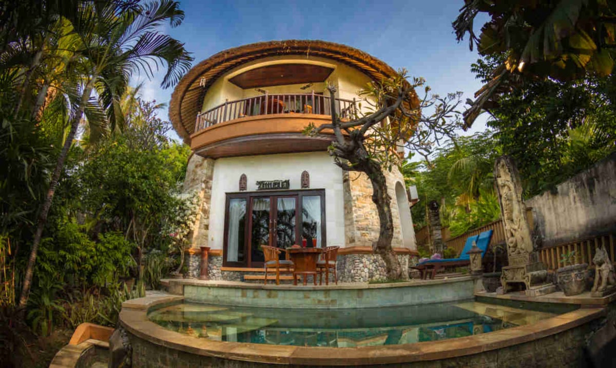 A unique round villa features a private pool surrounded by lush greenery. The structure's exterior is adorned with natural stone and wooden accents, emphasizing its rustic charm. A spacious balcony offers a view of the surroundings, with outdoor seating inviting relaxation.