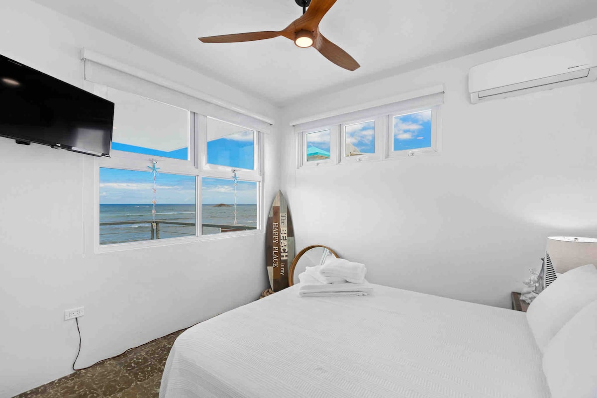 A bright bedroom features a spacious bed with fresh linens, accompanied by a decorative surfboard and a bedside lamp. Large windows provide views of the ocean and allow natural light to fill the room. A wall-mounted TV is positioned above a nightstand.