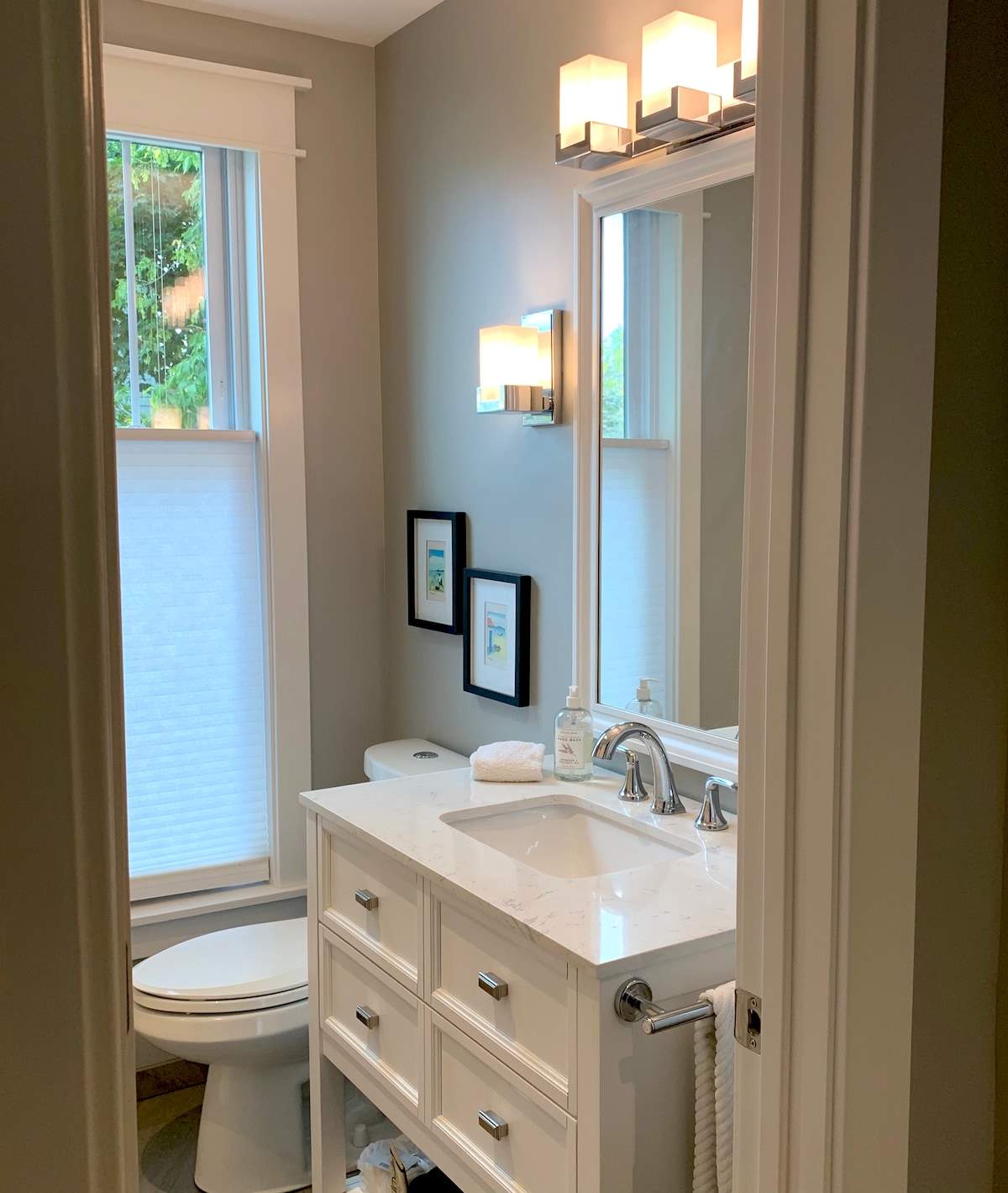 A well-lit bathroom offers a compact design with a white vanity topped with marble. Above, a mirror is flanked by modern lighting fixtures. A toilet is positioned adjacent to a window covered with sheer shades, and two framed artworks are displayed on the wall.