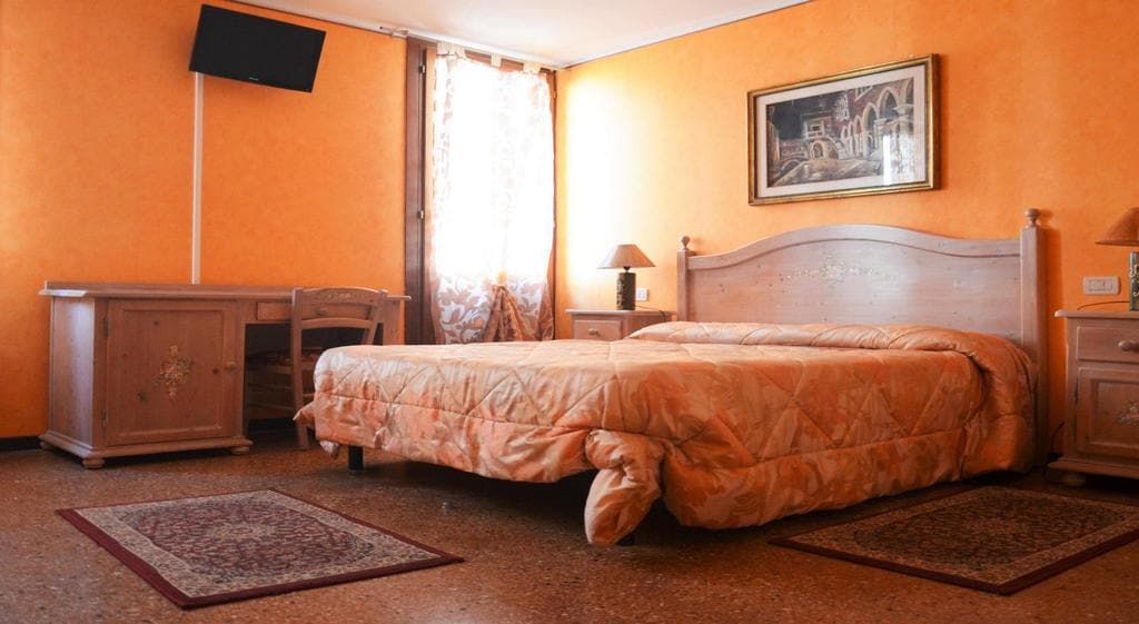 A cozy room features warm orange walls and a spacious bed with a patterned cover. Nearby, a desk and chair provide a workspace. Two lamps flank the bed, and a piece of wall art adds character. Natural light filters through the window dressed in light curtains.