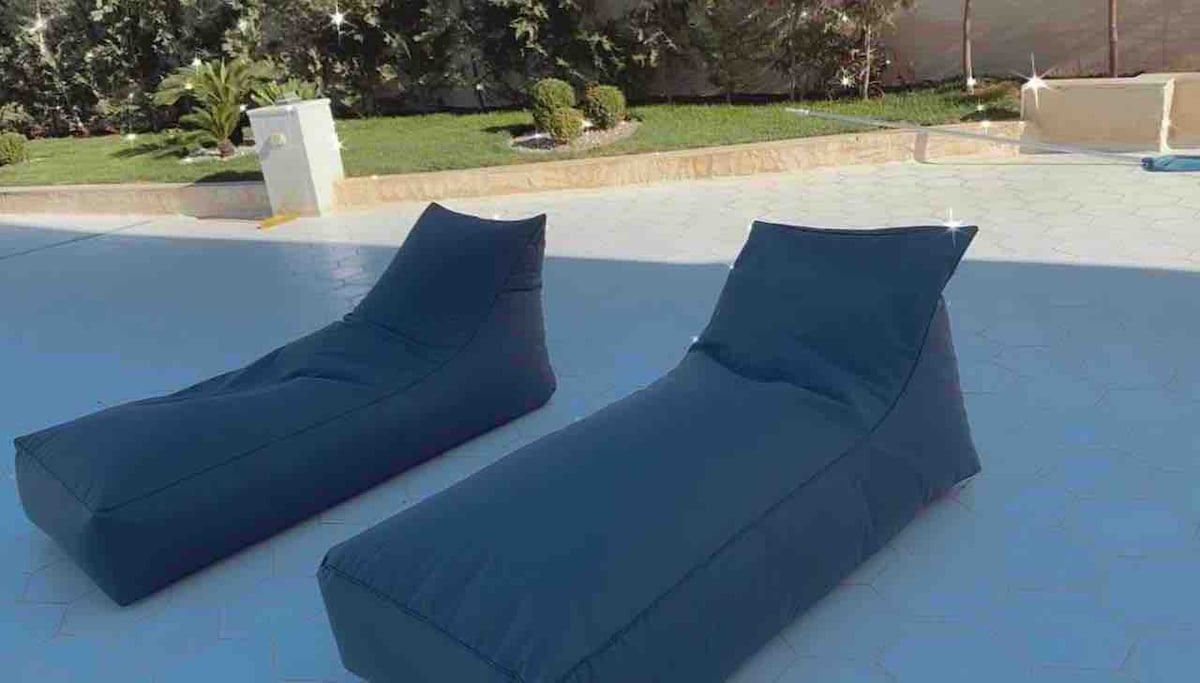 Two contemporary lounge chairs are positioned on a sunlit patio, surrounded by a landscaped garden. The chairs feature a modern design with soft, padded surfaces for relaxation. Greenery is visible in the background, enhancing the outdoor setting.
