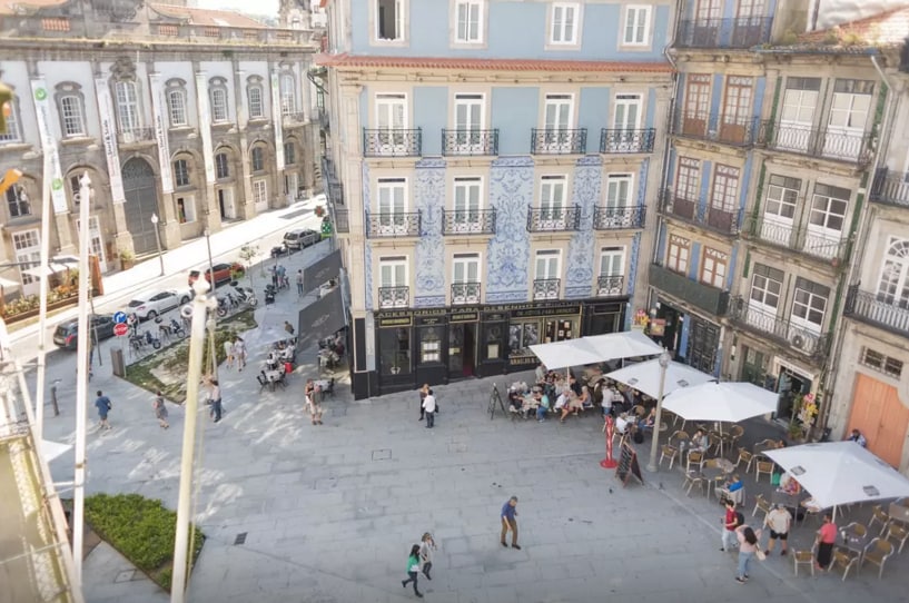 An aerial view captures a bustling square framed by traditional buildings adorned with blue tiles. Outdoor seating at a nearby café invites guests, while pedestrians stroll along the stone pavement. Trees and traffic signs create a vibrant urban scene in the heart of Porto.