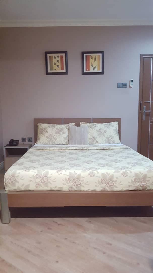 A private room is presented with a spacious bed adorned with a floral-patterned duvet and matching pillows. Two framed artworks are displayed above the bed, and a telephone is positioned nearby on a side table. The walls are painted in a subtle shade of pink.