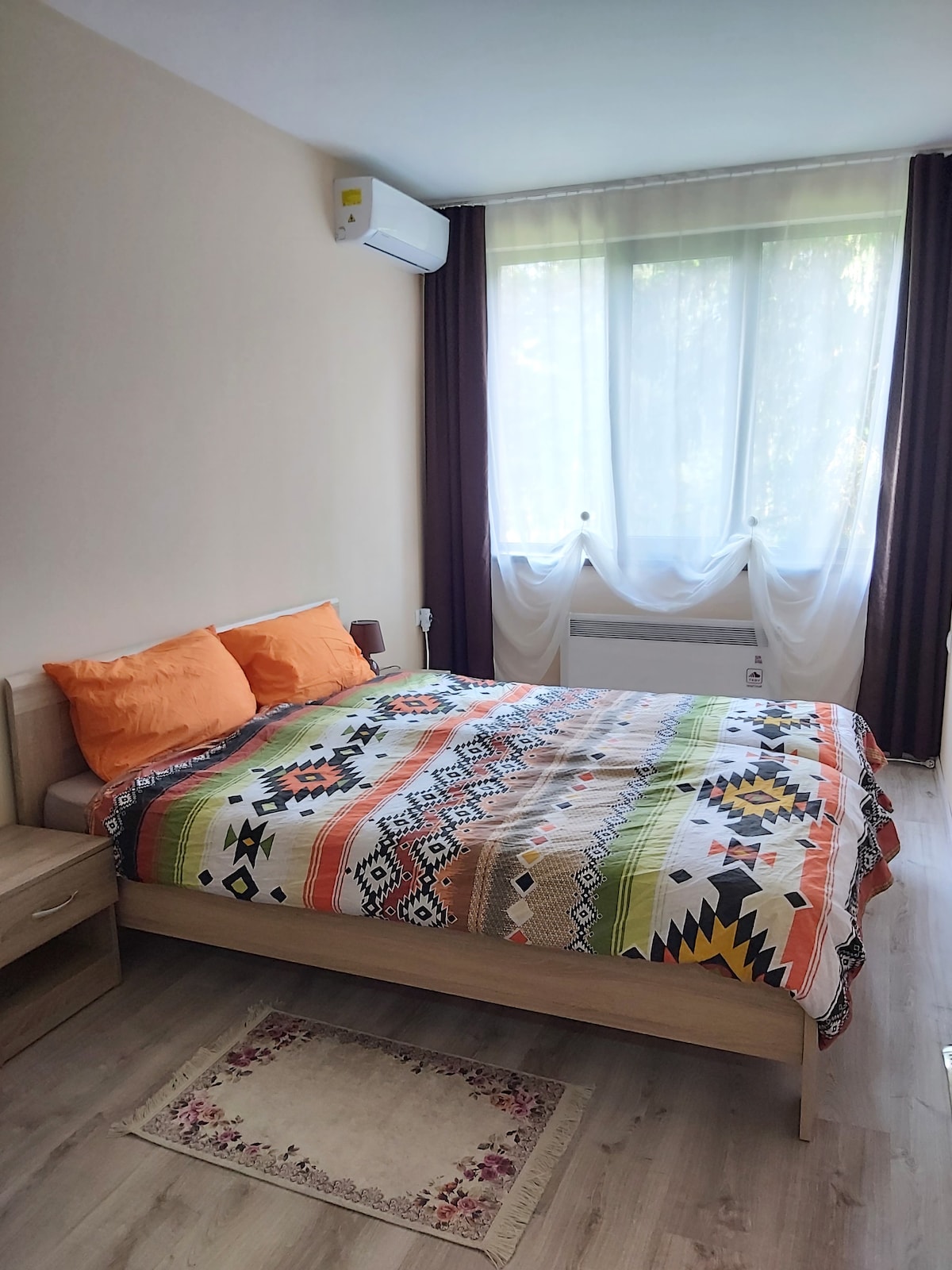 A bright sleeping area features a spacious bed with an orange and patterned blanket. Natural light enters through a large window, draped with sheer curtains. A wooden bedside table is positioned beside the bed, contributing to a functional and comfortable space.