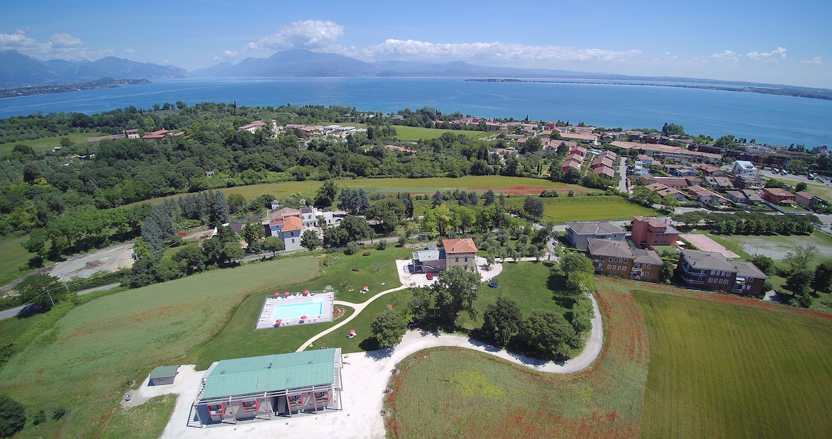 The aerial view captures the expansive landscape surrounding the property, with lush green fields and a vibrant blue lake in the background. The well-maintained grounds feature a swimming pool and a variety of trees, accentuating the serene environment.