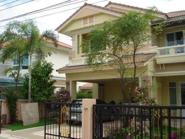 A two-story house showcases a beige facade complemented by a tiled roof. Lush tropical plants and small trees surround the structure, while a neatly maintained garden is visible. A gated driveway leads to a covered parking area under the house, enhancing the overall accessibility.