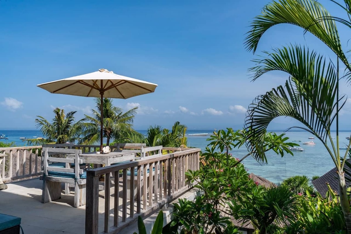 A spacious deck area is shown, featuring a large table and chairs shaded by a wide umbrella. Lush tropical plants surround the space, providing a natural ambiance. The view overlooks a calm turquoise sea, with boats gently bobbing in the distance under a clear blue sky.