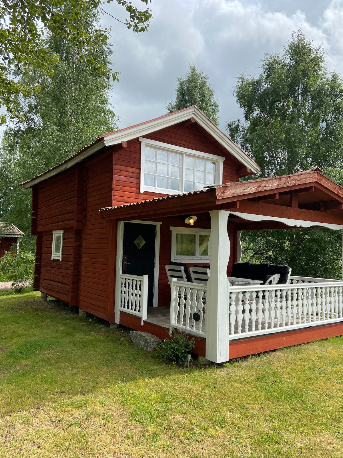 A charming two-story cabin with a bright red exterior features a covered porch and white railing. Comfortable seating is arranged on the porch, providing a welcoming space to relax. The surrounding green lawn and trees offer a natural setting, enhancing the overall ambiance.