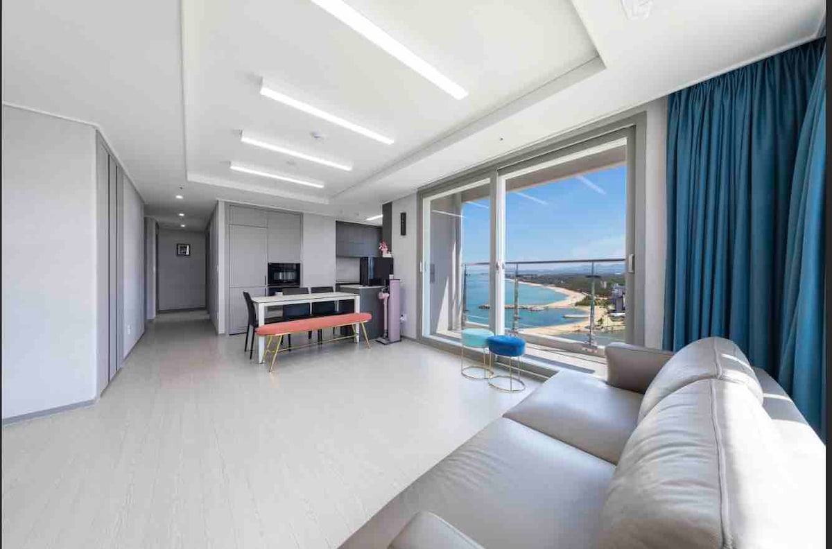 A bright and airy living space features large windows offering views of the coastline. A dining table is positioned near the kitchen, while a comfortable sofa invites relaxation. A pop of color is provided by a blue chair, enhancing the modern design.
