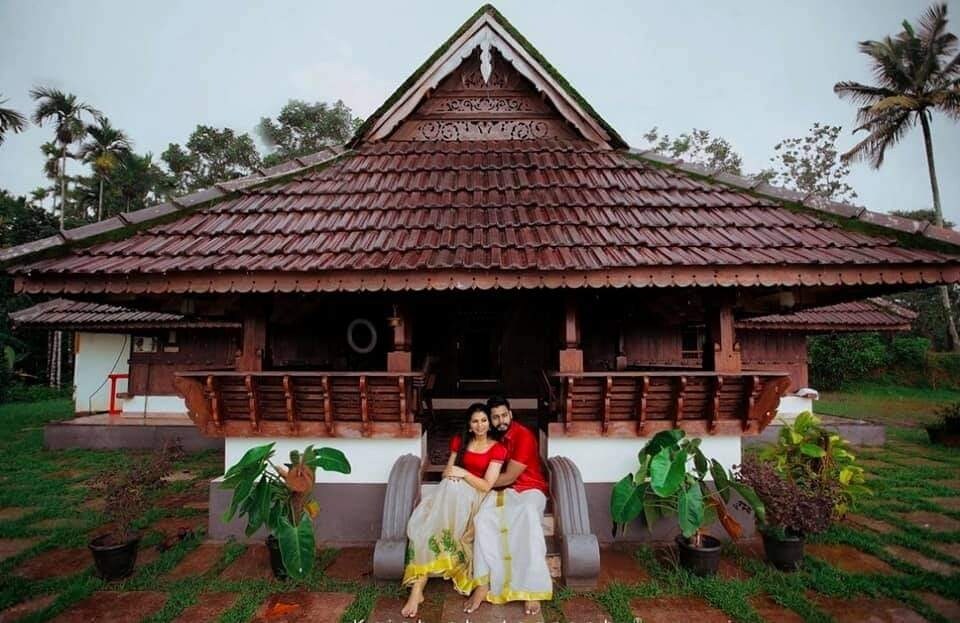 A traditional heritage home is displayed, showcasing intricate wooden architecture and a steep, tiled roof. Lush greenery surrounds the property, and a couple is seated on a bench, contributing to a serene ambience. The building offers sheltered outdoor space, ideal for relaxation.