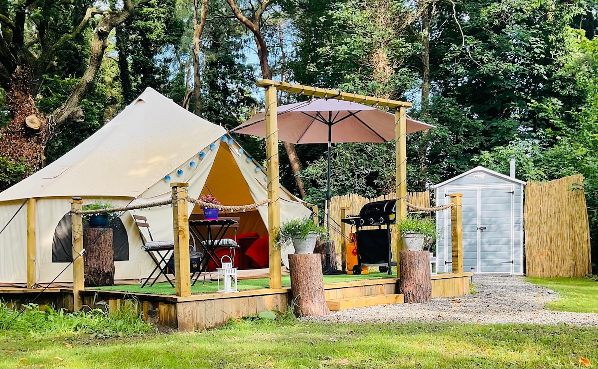 A spacious tent is set within a private wooded area, featuring a wooden deck with an umbrella and barbecue. Potted plants are placed on the deck, and a small shed is visible in the background, framed by lush greenery.