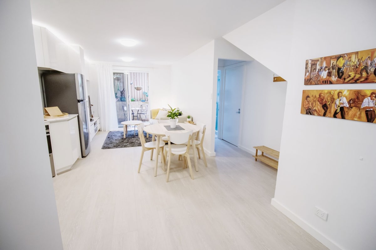 An open-plan living space is displayed, featuring a round dining table surrounded by light-colored chairs. A modern kitchen area with sleek cabinetry and stainless-steel appliances is visible. Natural light fills the room, enhancing the clean, airy atmosphere. A door leads to an outdoor patio.