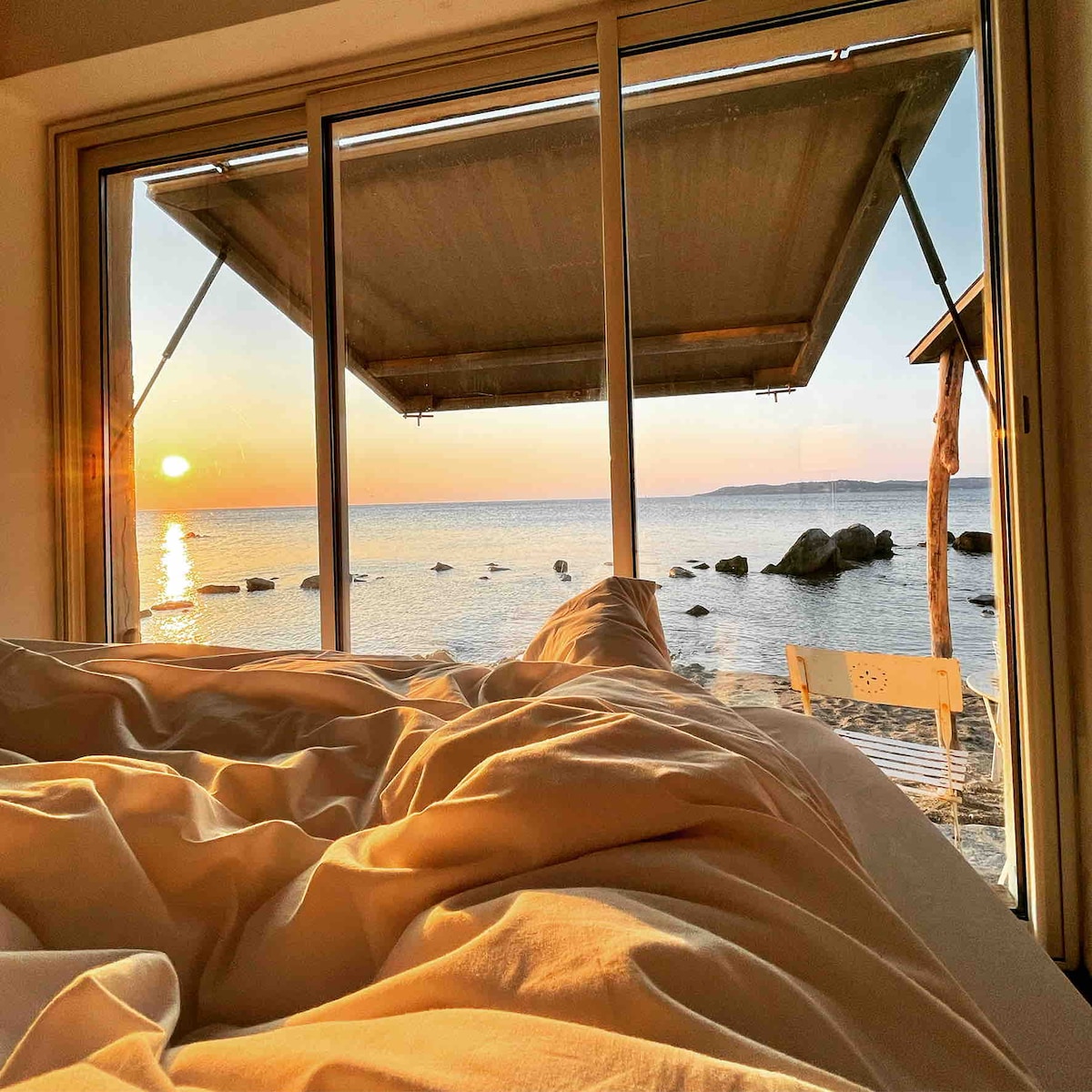 A spacious bed is framed by large glass doors, revealing a serene view of the ocean at sunset. Soft, light-colored bedding is arranged casually. The scene is complemented by the soft glow of the setting sun and the tranquil water visible beyond the bed.