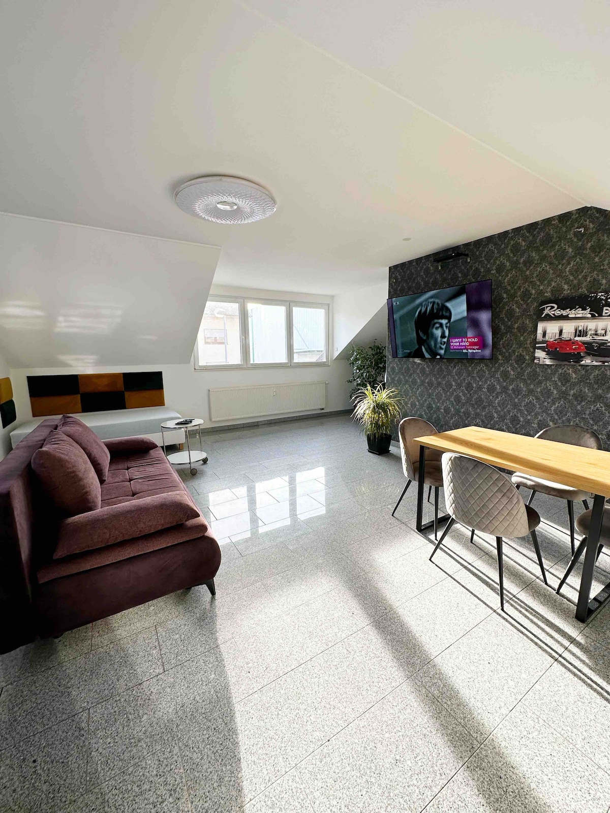 A spacious living area features a plush sofa and a dining table with stylish chairs. Natural light floods through large windows, illuminating the sleek flooring. A flat-screen TV is mounted on one wall, complementing the contemporary decor.