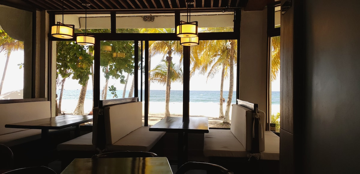 An inviting dining area features several booths and tables arranged to maximize views of the ocean. Large windows frame the scene, offering glimpses of palm trees and the shoreline beyond while natural light fills the space.