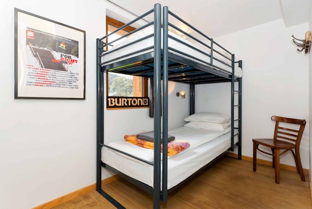 A cozy bedroom features a metal bunk bed with two levels of mattresses. A chair is positioned nearby, and a wall-mounted light fixture is visible. Natural light enters through a window, illuminating the wooden flooring and framed artwork on the wall.
