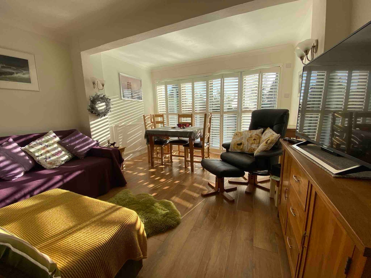 An open plan sitting area features a comfortable sofa and a reclining chair beside a wooden table and dining chairs. Light filters through wooden slatted shutters, creating a warm ambiance. A television is positioned on a wooden cabinet, complemented by soft furnishings in calming colors.