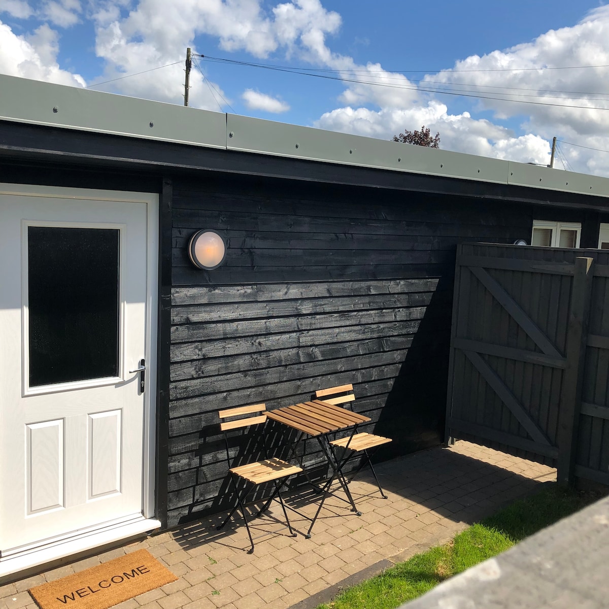 A compact outdoor seating area features a small table accompanied by two chairs. The cabin's entrance is highlighted by a light fixture beside the door. A welcome mat is positioned in front of the entrance, and a partially fenced area is visible, enhancing privacy.