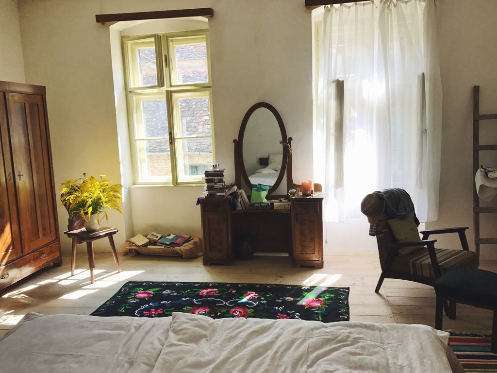 A welcoming bedroom features a spacious bed with light bedding and a decorative rug. A vintage wardrobe stands beside a table with a vibrant flower arrangement. Sunlight filters through sheer curtains, illuminating a dresser with reflective surfaces and a chair that enhances the room's cozy ambiance.