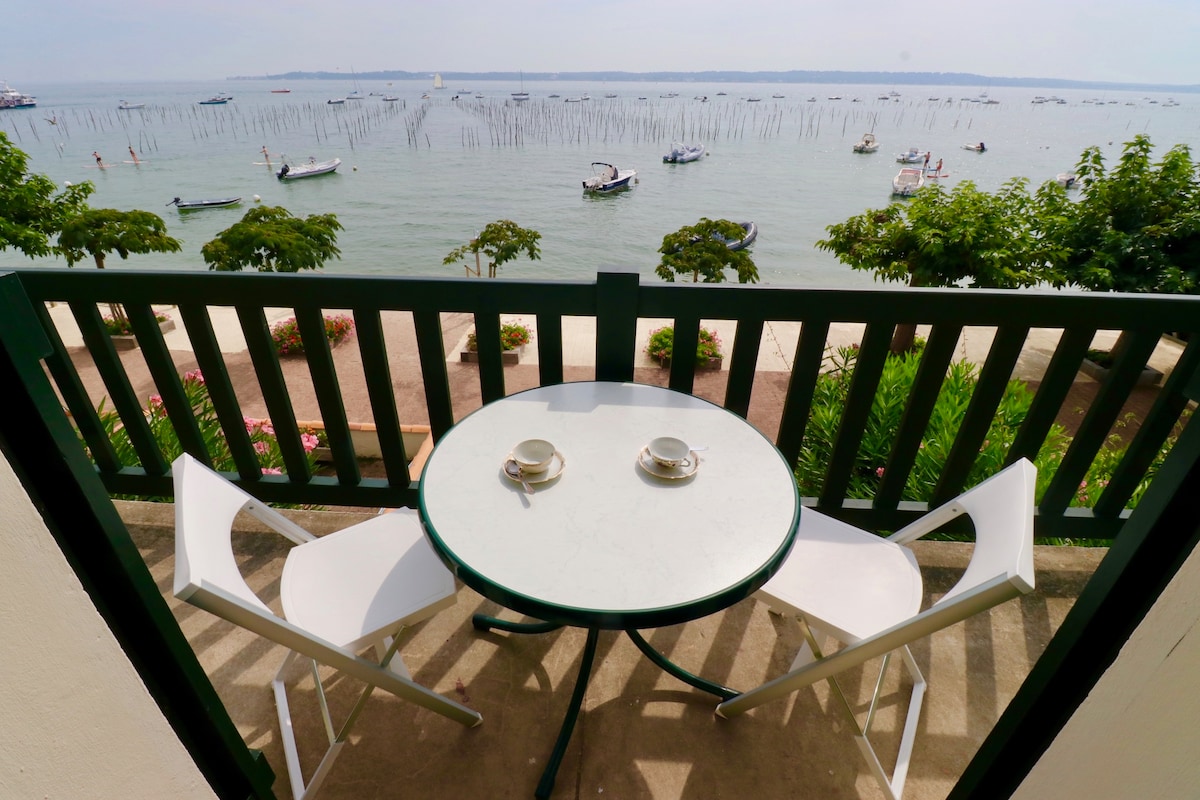 A small balcony is presented with a round table and two white chairs, overlooking the bay. Boats float in the water, while greenery frames the scene. The calm atmosphere invites relaxation, complemented by a view of the sky and distant horizons.