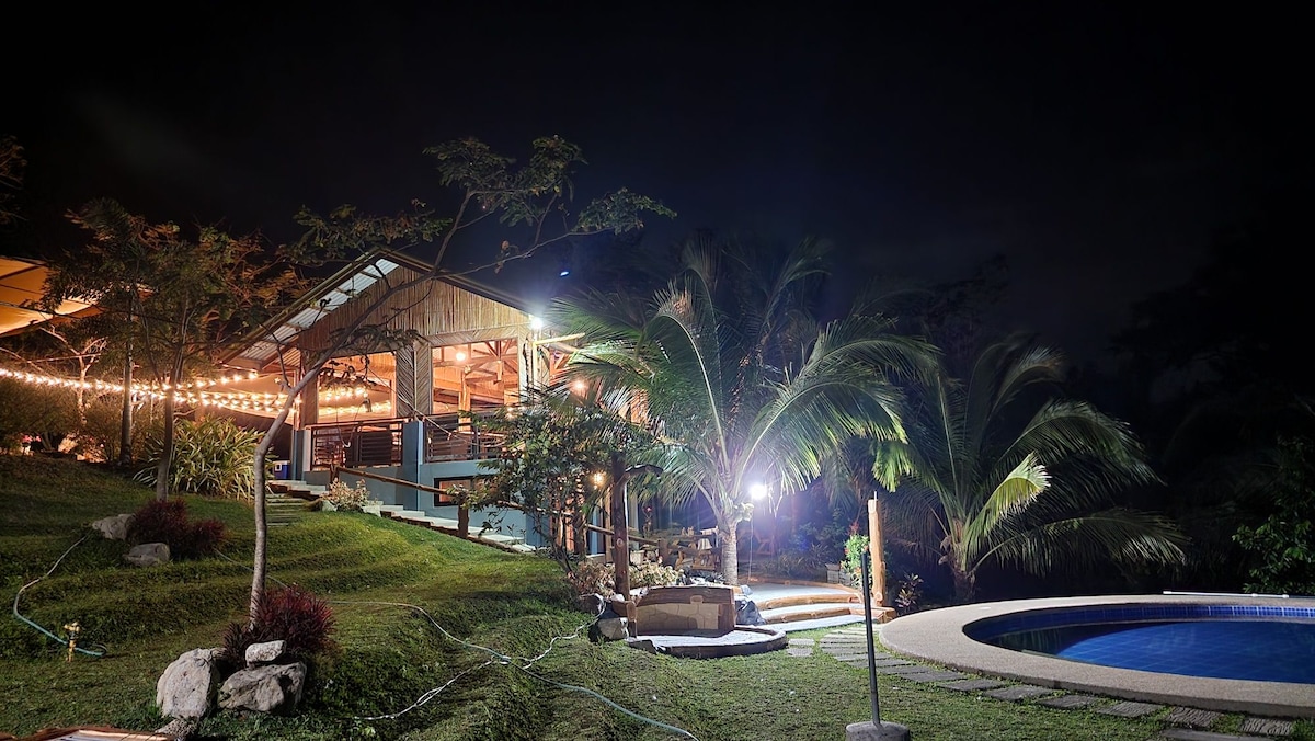 A serene outdoor space is illuminated by soft lighting, highlighting a cozy gathering area adjacent to the pool. Lush greenery surrounds the area, with palm trees swaying gently in the breeze, creating a peaceful ambiance under the night sky.