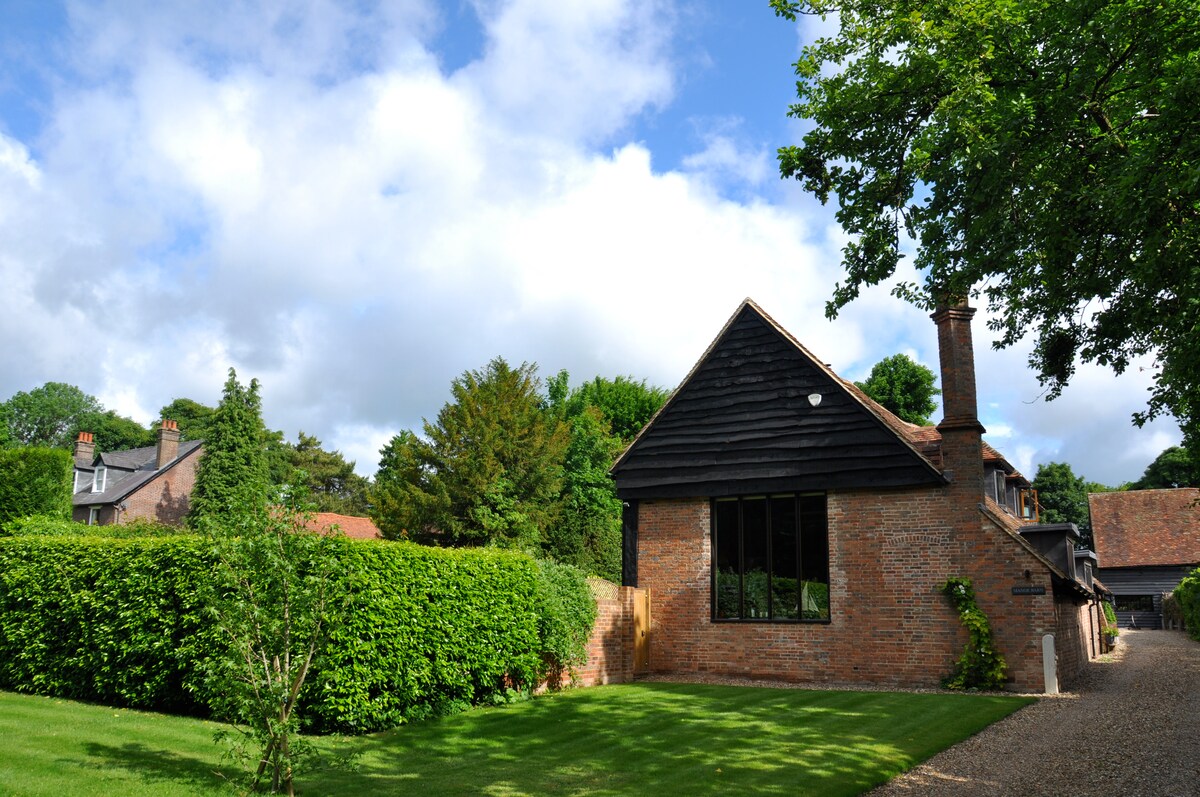 A well-maintained exterior of the Lodge is showcased, featuring a blend of brick and timber elements under a partly cloudy sky. Surrounding greenery and neatly trimmed hedges provide a serene atmosphere, while a gravel pathway leads to the entrance, enhancing the inviting setting.