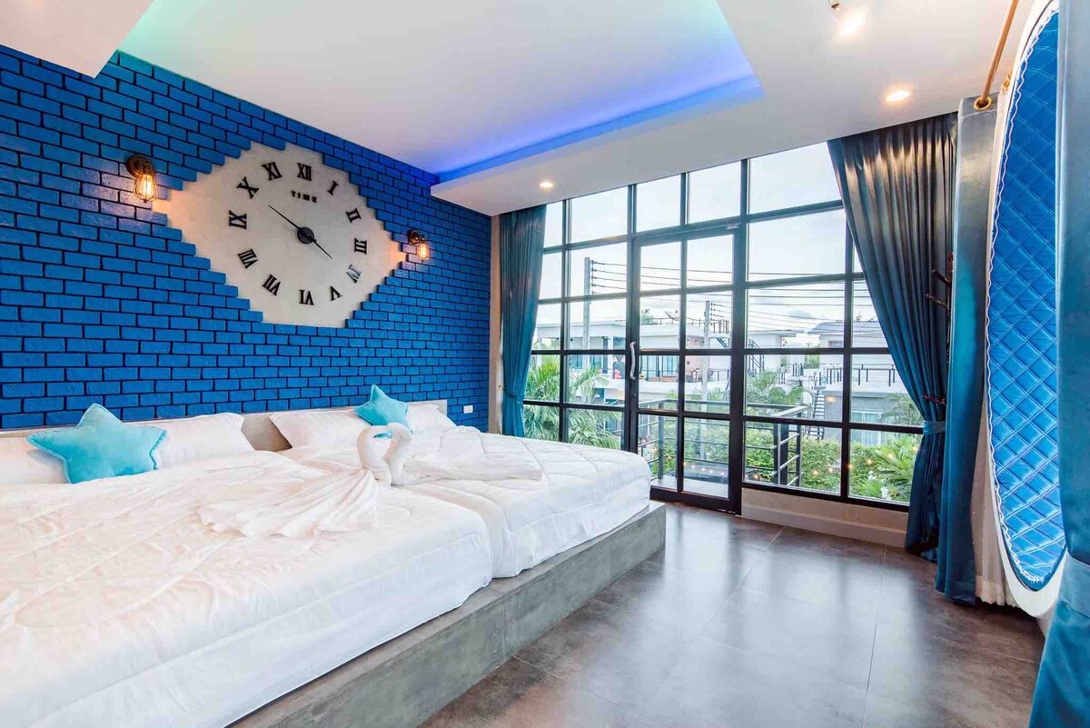 A bright bedroom features large windows with views of the surrounding area, allowing natural light to fill the space. Two spacious beds are arranged on a sleek platform. The blue accent wall is complemented by contemporary wall lights and decorative elements that enhance the room's ambiance.