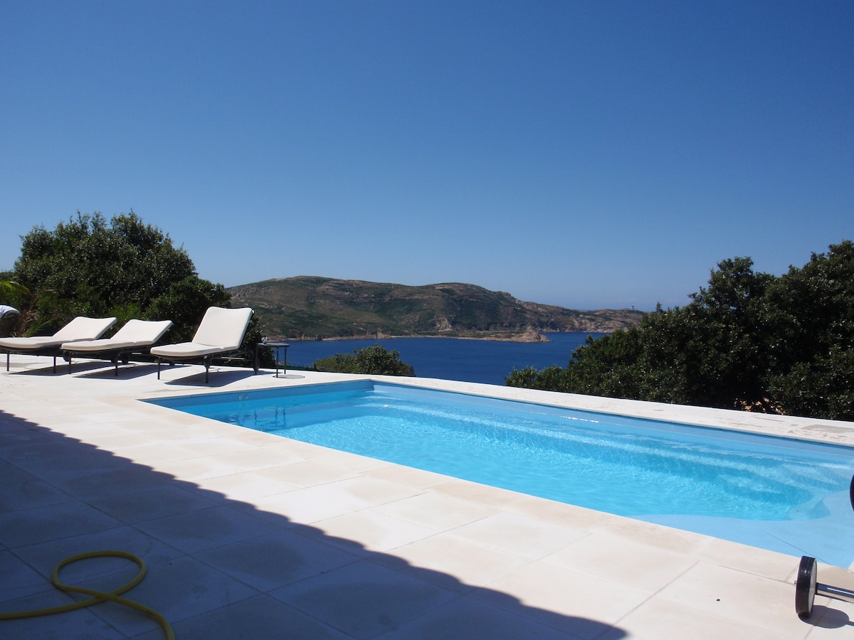 A serene scene is displayed with a heated swimming pool reflecting clear blue skies. Comfortable lounge chairs are positioned alongside the pool, offering a view of the expansive sea and distant hills, surrounded by lush greenery for a tranquil setting.