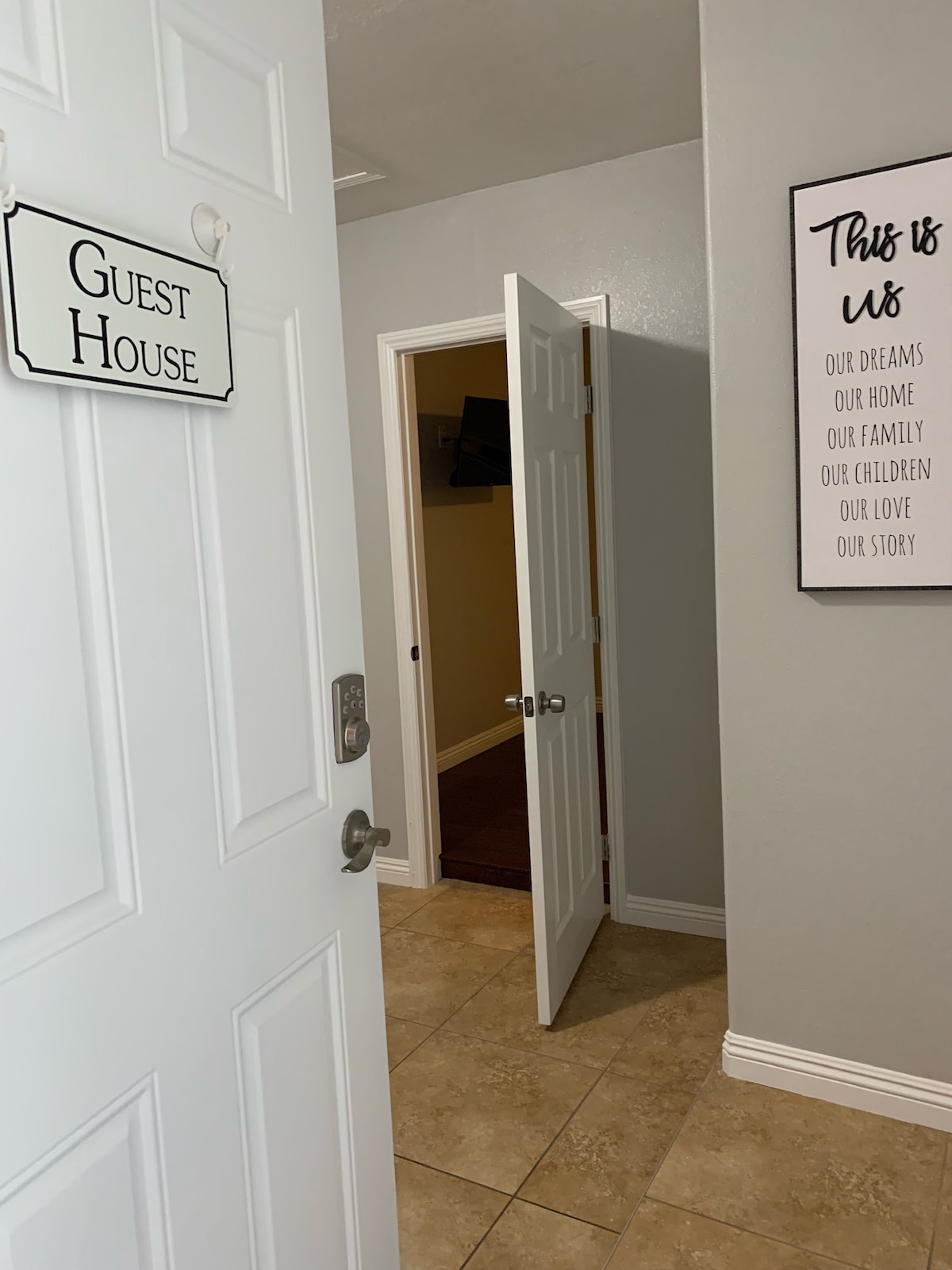 The entryway to the guest house features a white door with a sign reading 'Guest House.' A light gray wall complements beige tiled flooring. An open interior door is visible in the background, leading into a warmly lit room that includes a wall-mounted television.