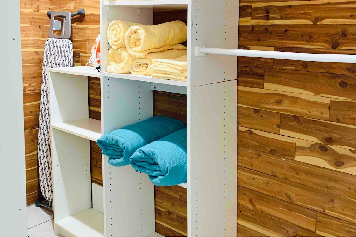A neatly organized storage area features shelves holding neatly stacked yellow towels and blue bed covers. An ironing board is positioned beside the storage unit, with a light brown wooden wall serving as a backdrop.