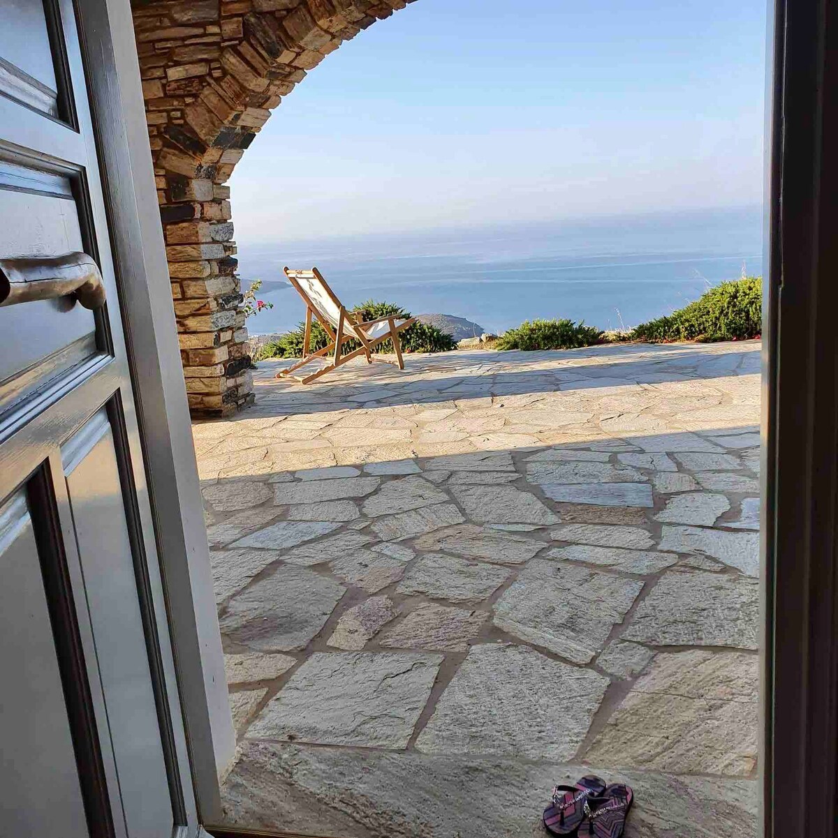 A stone entrance reveals a spacious outdoor area, featuring a wooden deck chair and a stunning view of the deep blue sea. The ground is paved with large stone slabs, while a pair of sandals rests just inside the doorway, adding a touch of casual comfort.