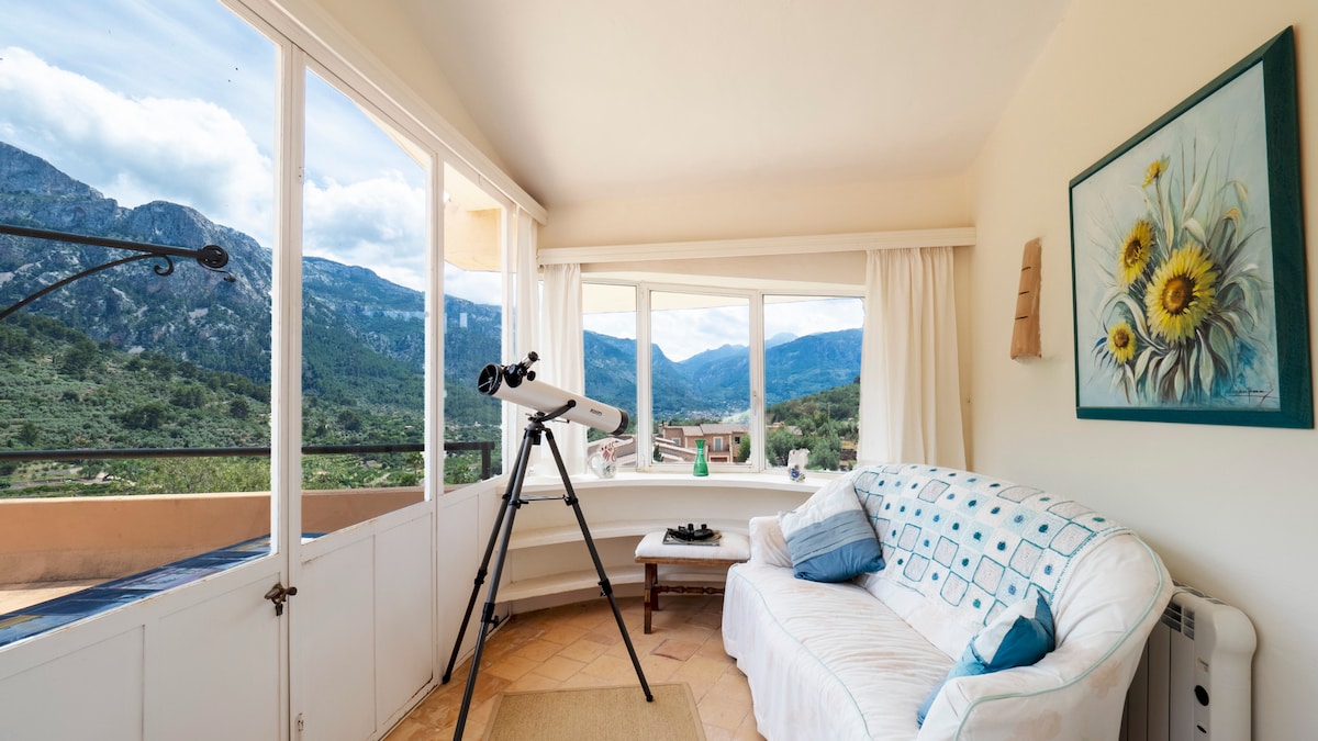 A bright living area features a comfortable white sofa, complemented by a decorative piece with sunflowers. A telescope is positioned near floor-to-ceiling windows, providing captivating views of the surrounding mountains and lush landscape, enhancing the light-filled space.