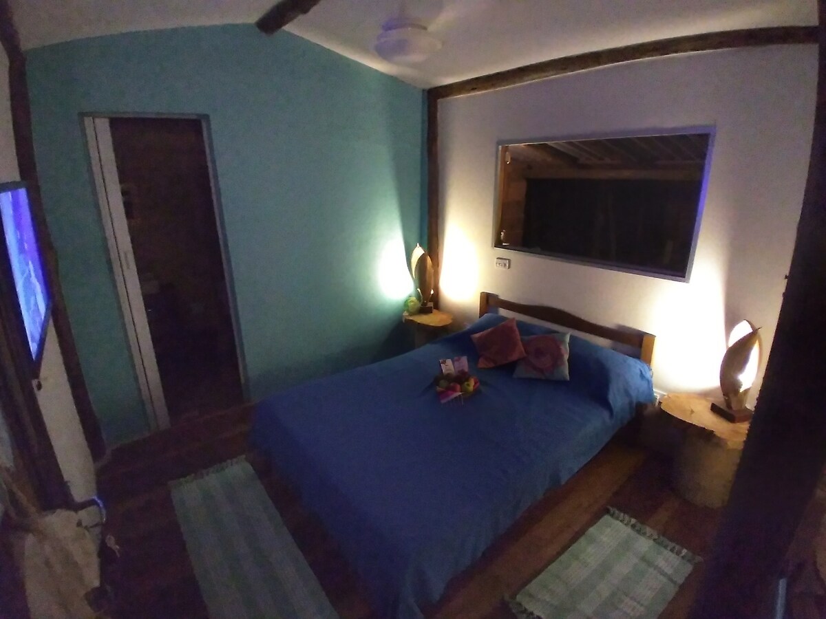 A tranquil bedroom features a spacious bed topped with a blue coverlet. Soft lighting is provided by two bedside lamps, complementing the vibrant blue walls. A large window offers a view, while textured rugs and wooden accents enhance the serene setting.