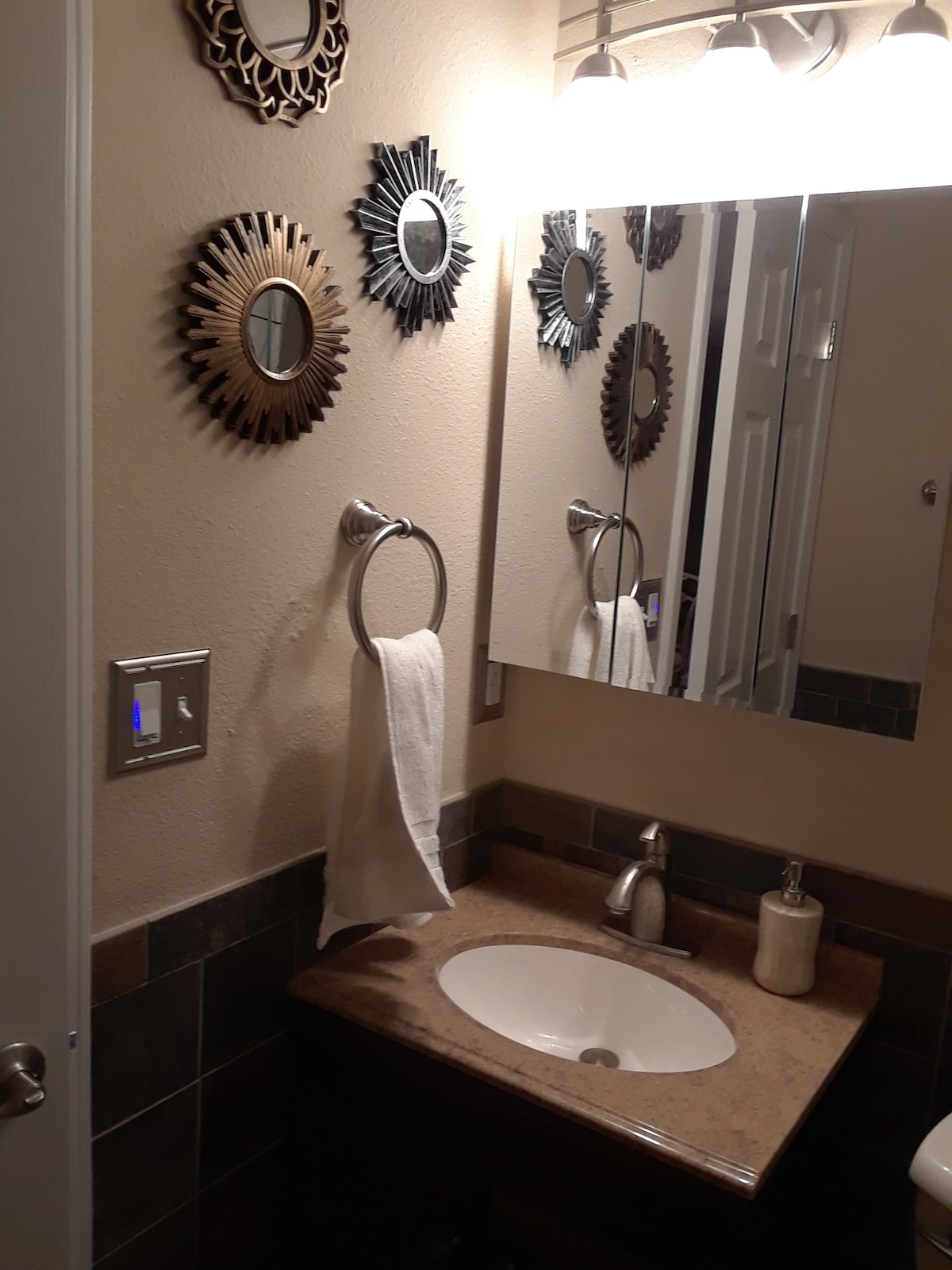 The bathroom features a sleek vanity with a round sink and countertop. Mirrors with decorative frames enhance the wall. A white towel hangs on a polished towel ring, and a soap dispenser is positioned beside the sink. Soft lighting creates a warm ambiance.
