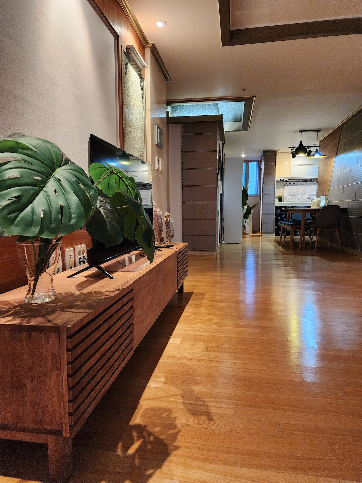 A hallway is presented with polished wooden flooring, leading toward a cozy dining area in the background. A wooden console features decorative plants and sculptures, while soft lighting enhances the inviting atmosphere of the space.